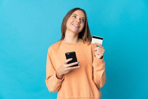 Young Lithuanian woman isolated on blue background buying with the mobile with a credit card while thinking