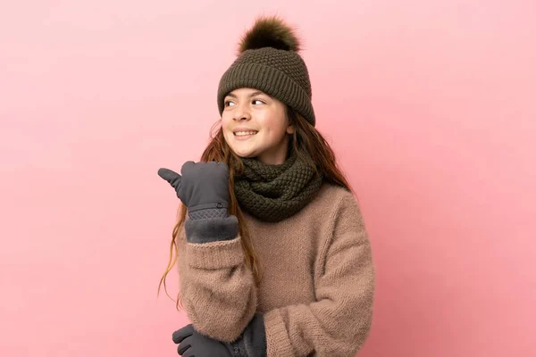 一个戴着冬帽的小女孩 被粉色背景隔离在一边 指着侧面展示自己的产品 — 图库照片