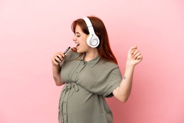 Genç kızıl saçlı beyaz kadın pembe arka planda izole edilmiş hamile ve müzik dinlerken dans ediyor.