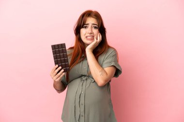 Genç kızıl saçlı beyaz kadın pembe arka planda izole edilmiş hamile ve elinde aç çikolatayla.