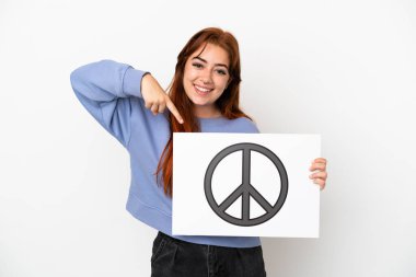 Genç kızıl saçlı kadın beyaz arka planda izole edilmiş. Elinde barış sembolü olan bir pankart var ve onu işaret ediyor.