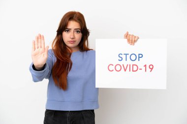 Beyaz arka planda izole edilmiş genç kızıl saçlı bir kadın elinde yazılı bir pankartla Covid 19 'u durdurun ve dur işareti yapıyor.