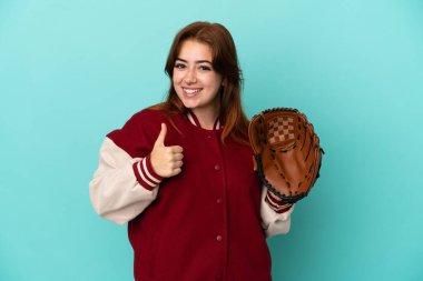 Beyzbol oynayan genç kızıl saçlı kadın mavi arka planda izole edilmiş bir şekilde baş parmak hareketi yapıyor.