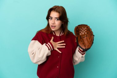 Beyzbol oynayan genç kızıl saçlı kadın mavi arka planda izole edilmiş, sağa bakarken şaşırmış ve şok olmuştu.