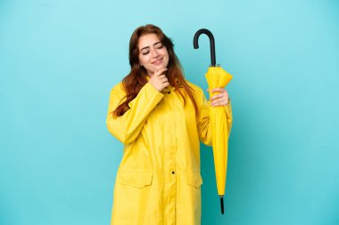 Mavi arka planda şemsiyesi olan kızıl saçlı kadın yana bakıyor ve gülümsüyor.