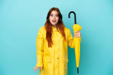 Mavi arka planda şemsiyesi olan kızıl saçlı kadın sürpriz bir yüz ifadesiyle izole edilmiş.