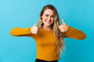 Mavi arka planda izole edilmiş genç Brezilyalı kadın baş parmak hareketi yapıyor.