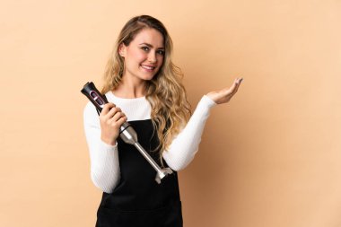 Bej arka planda izole edilmiş el blenderi kullanan genç Brezilyalı kadın davet etmek için ellerini yana uzatıyor.