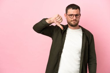 Pembe arka planda izole edilmiş genç Brezilyalı adam olumsuz ifadeyle baş parmağını aşağı gösteriyor.