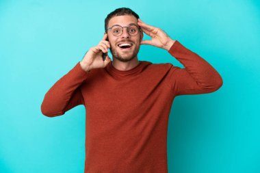 Mavi arka planda izole edilmiş cep telefonu kullanan Brezilyalı genç adam sürpriz bir ifadeyle
