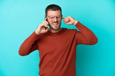 Mavi arka planda izole edilmiş cep telefonu kullanan Brezilyalı genç adam hayal kırıklığına uğramış ve kulaklarını kapatmış.