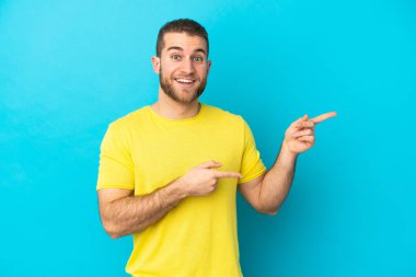 Mavi arka planda izole edilmiş yakışıklı beyaz adam şaşırmış ve işaret ediyor.