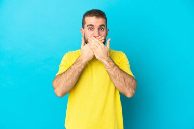 Mavi arka planda izole edilmiş yakışıklı beyaz adam elleriyle ağzını kapatıyor.
