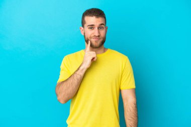 Genç yakışıklı beyaz adam mavi arka planda izole edilmiş ve düşünüyor.