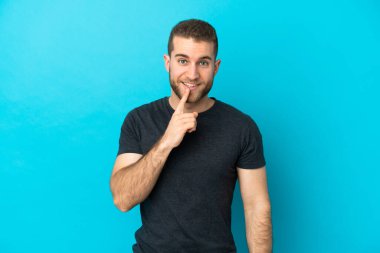 Mavi arka planda izole edilmiş yakışıklı beyaz adam sessiz bir jest işareti gösteriyor. Parmağını ağzına sokuyor.