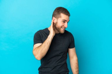 Genç beyaz adam mavi arka planda izole edilmiş bir şekilde elini kulağına koyarak bir şeyler dinliyor.