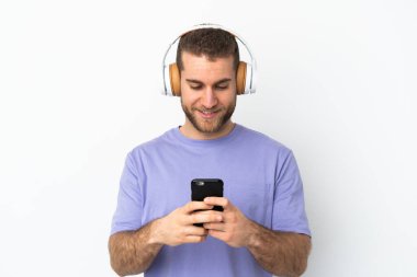 Beyaz arka planda izole edilmiş yakışıklı beyaz adam müzik dinliyor ve cep telefonu arıyor.