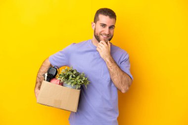 Genç Kafkasyalı, sarı arka planda izole edilmiş eşyalarla dolu bir kutuyu kaldırırken hamle yapıyor ve gülümsüyor.