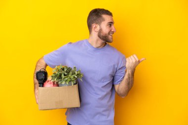 Genç Kafkasyalı, sarı arka planda izole edilmiş eşyalarla dolu bir kutuyu kaldırırken bir ürün sunmak için yan tarafı işaret ediyor.