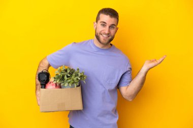 Genç Kafkasyalı, sarı arka planda izole edilmiş eşyalarla dolu bir kutuyu kaldırırken hareket ediyor. Davet etmek için ellerini yana açıyor.