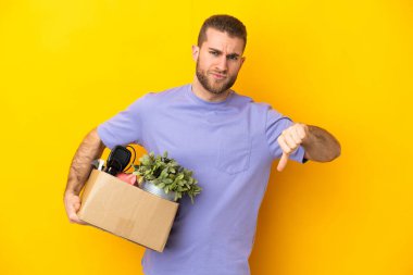 Genç Kafkasyalı, sarı arka planda izole edilmiş şeylerle dolu bir kutuyu seçerken negatif ifadeyle baş parmağını aşağı gösteriyor.