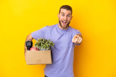 Genç Kafkasyalı, sarı arka planda izole edilmiş eşyalarla dolu bir kutuyu alırken şaşırdı ve ön tarafı işaret etti.