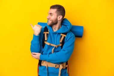 Sarı arka planda izole edilmiş büyük sırt çantalı beyaz bir dağcı bir ürünü sunmak için kenarı işaret ediyor.