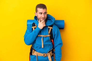 Sarı arka planda izole edilmiş büyük bir sırt çantası olan beyaz dağcı yan tarafa bakıyor ve gülümsüyor.
