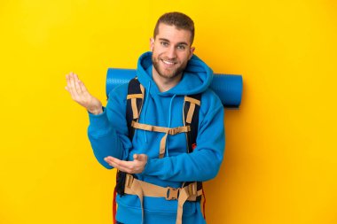 Sarı arka planda izole edilmiş büyük bir sırt çantası olan beyaz, genç bir dağcı davet edildiği için elini yana uzatıyor.