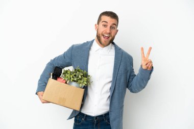 Beyaz arka planda izole edilmiş şeylerle dolu bir kutuyu alırken gülümseyen ve zafer işareti gösteren genç bir Kafkasyalı hamle yapıyor.