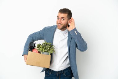 Beyaz arka planda izole edilmiş eşyalarla dolu bir kutuyu alıp elini kulağına koyarak bir şey dinleyen genç beyaz bir adam hamle yapıyor.