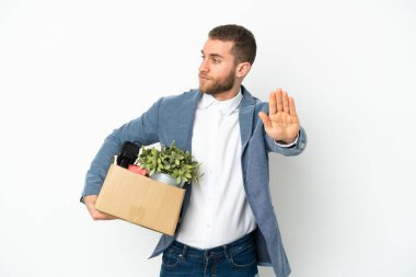 Beyaz arka planda izole edilmiş eşyalarla dolu bir kutuyu kaldırırken hareket eden genç beyaz adam dur hareketi yapıyor ve hayal kırıklığına uğruyor.