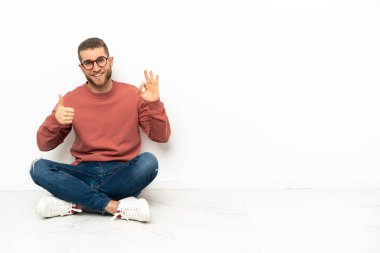 Yerde oturan yakışıklı genç adam işaret veriyor ve başparmağını kaldırıyor.