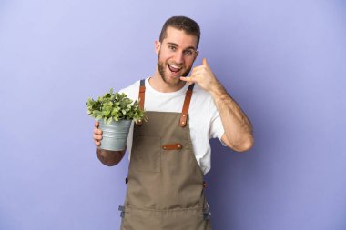 Bahçıvan beyaz bir adam sarı arka planda izole edilmiş bir bitki tutuyor ve telefon jesti yapıyor. Bana geri dönüş işareti ver.