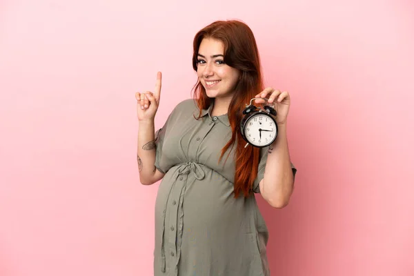 若いです赤毛Caucasian女性はピンクの背景妊娠中や時計を保持しながら 1をカウント — ストック写真