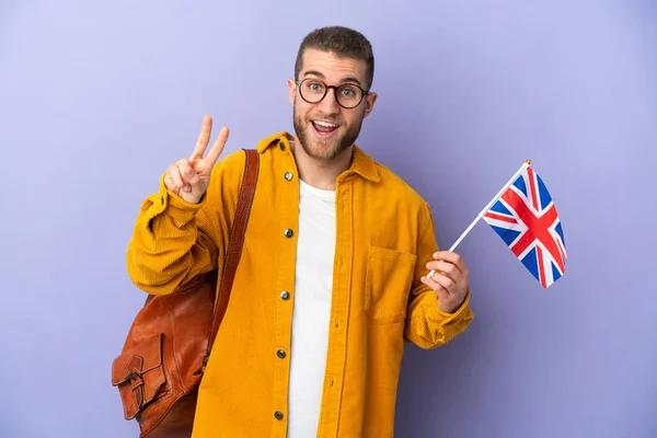 年轻的高加索男子举着一面英国国旗 与紫色背景隔离 面带微笑 并展示胜利的标志 — 图库照片