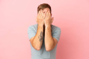 Kırmızımsı beyaz adam pembe arka planda izole edilmiş gözlerini elleriyle kapatıyor.