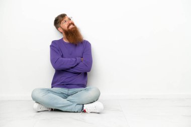 Beyaz arka planda izole bir şekilde yerde oturan beyaz tenli genç adam gülümserken yukarı bakıyor.