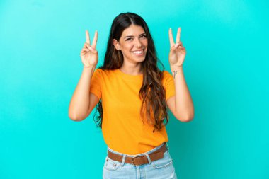 Mavi arka planda izole edilmiş genç beyaz kadın iki eliyle zafer işareti gösteriyor.
