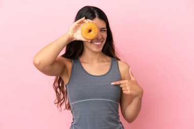 Pembe arka planda izole edilmiş genç beyaz kadın elinde donut ve mutlu.