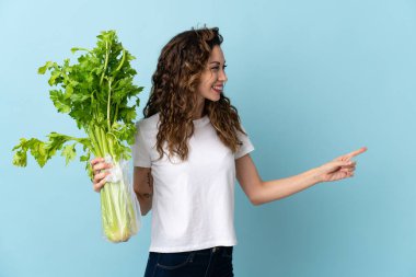 Mavi arka planda izole edilmiş bir kereviz tutan genç bir kadın bir ürünü sunmak için yan tarafı işaret ediyor.