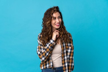 Mavi arka planda izole edilmiş genç beyaz kadın gülümserken yukarı bakıyor.