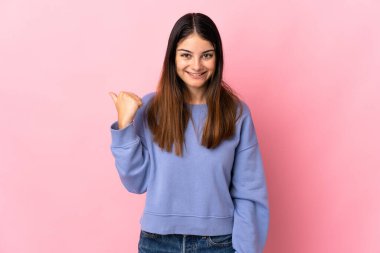 Pembe arka planda izole edilmiş genç beyaz kadın bir ürünü sunmak için kenarı işaret ediyor.