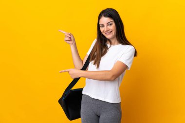 Sarı arka planda izole edilmiş spor çantalı genç bir kadın. Parmakla göstererek bir ürün sunuyor.