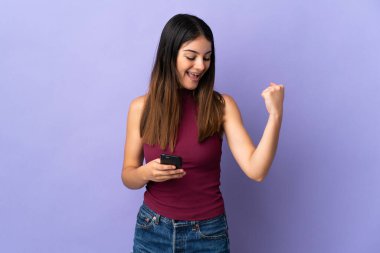 Mor arka planda izole edilmiş genç beyaz bir kadın ve telefon zafer pozisyonunda.