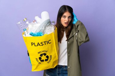 Elinde plastik şişelerle dolu bir çanta tutan genç bir kadın. Mor arka planda izole edilmiş, sürpriz bir ifadeyle geri dönüşüm için.