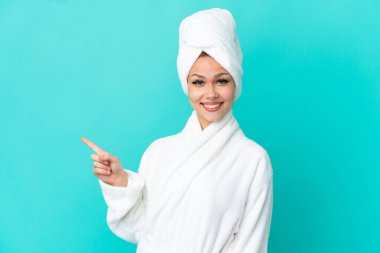 İzole edilmiş mavi arka planda bornozlu genç sarışın kız, parmağını yana doğru gösteriyor.