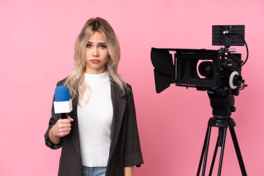 Elinde mikrofon tutan ve üzgün ve depresif bir ifadeyle pembe arka planda haber sunan gazeteci kadın.