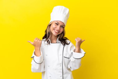 Sarı arka planda izole edilmiş genç bir Rus şef kız. Baş parmakları havada ve gülümsüyor.