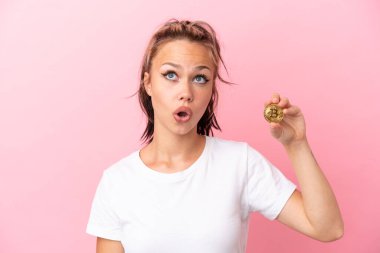 Elinde Bitcoin olan genç bir Rus kız pembe arka planda izole edilmiş yukarı bakıyor ve şaşırmış bir ifadeyle